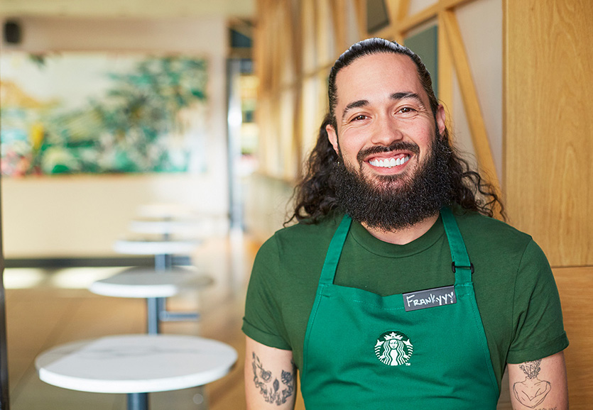 Une personne portant un tablier vert Starbucks sourit en regardant la caméra.