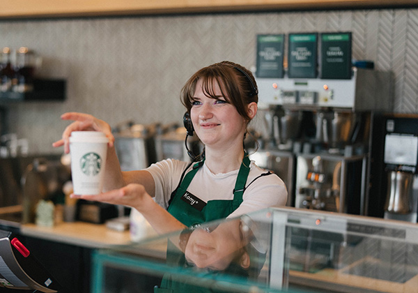 Une personne portant un tablier vert Starbucks présente en souriant une tasse blanche réutilisable Starbucks, le logo bien visible.