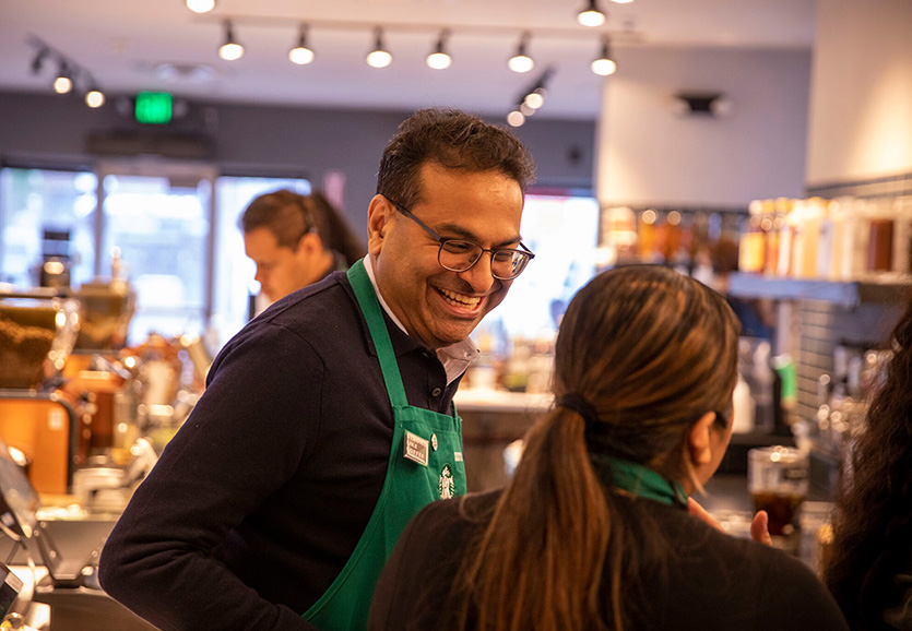 Le chef de la direction de Starbucks, Laxman Narasimhan, porte un tablier vert Starbucks et rigole avec une barista en travaillant dans un magasin.