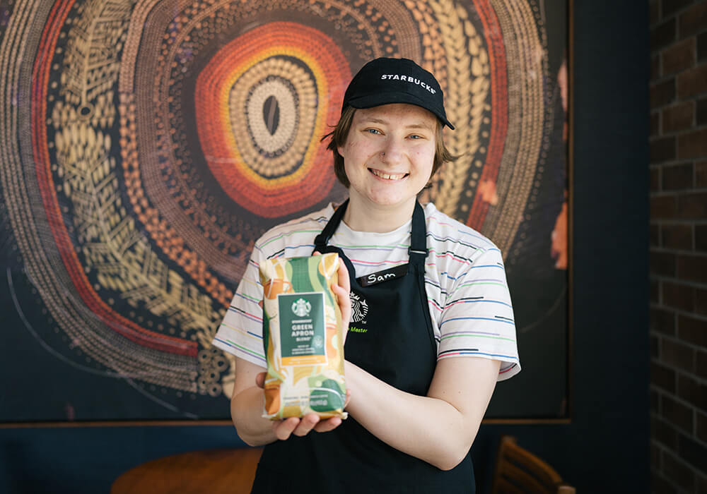 Une personne souriante portant un tablier noir Starbucks tient un paquet de café Mélange Tablier vert.
