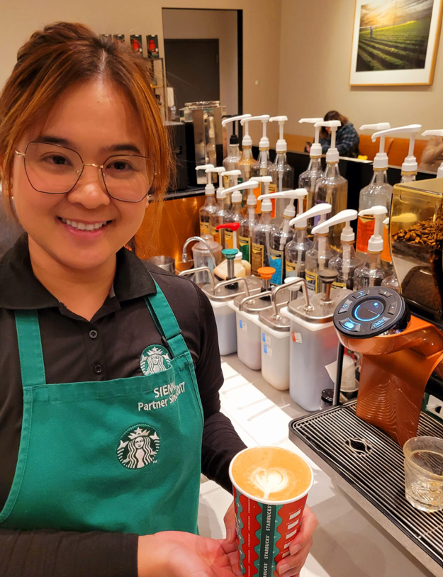 Une employée de Starbucks portant des lunettes et un tablier vert Starbucks, tient une tasse remplie d’une boisson garnie d’un motif d'art latte.