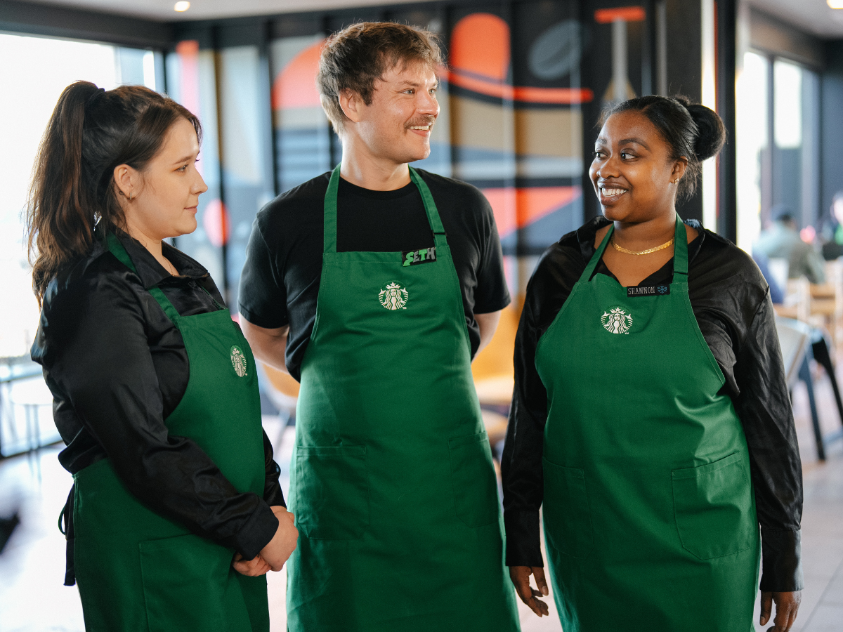 Trois personnes portant un tablier vert sourient en se tenant l’une à côté de l’autre dans un magasin Starbucks.