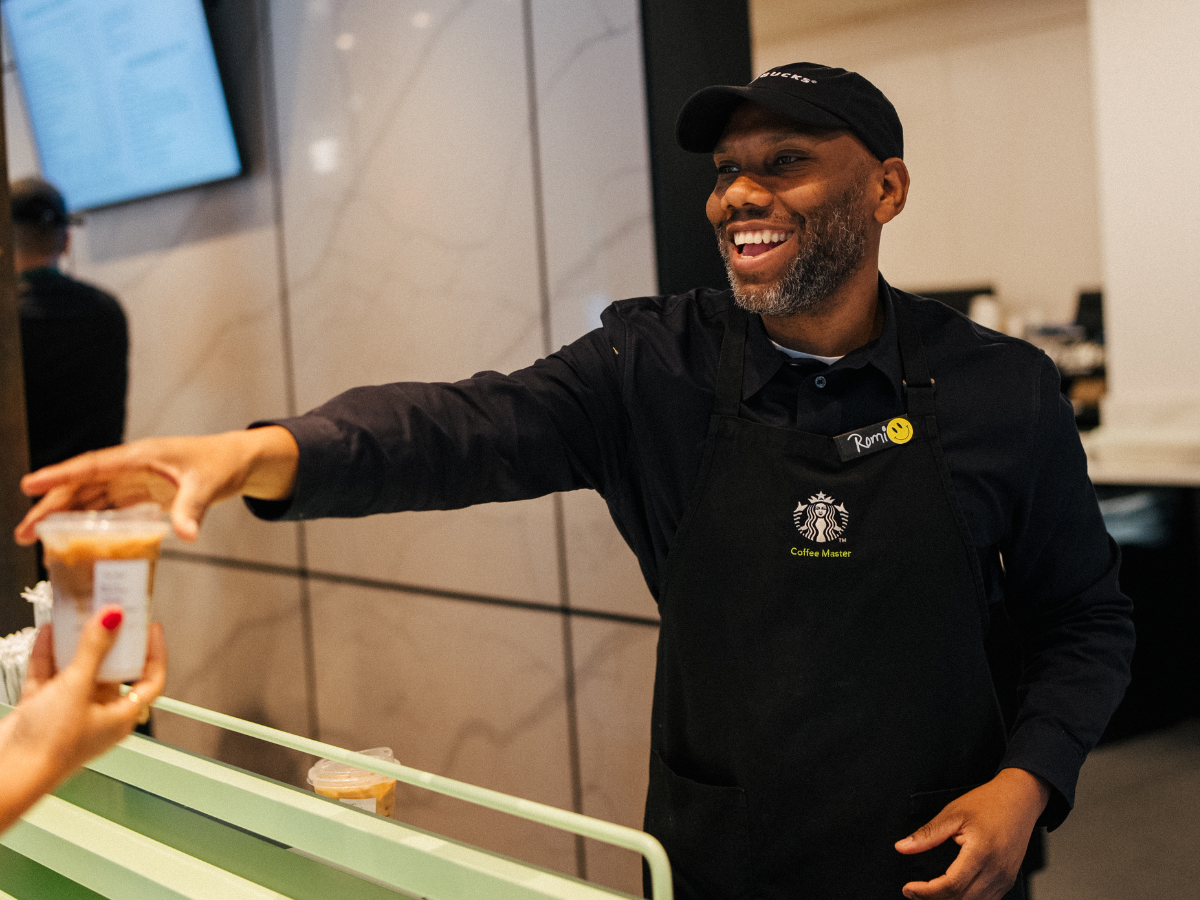  Une personne portant un tablier noir de maître de café Starbucks sourit en tendant un café à une cliente par-dessus le comptoir pendant qu’elle travaille chez Starbucks.