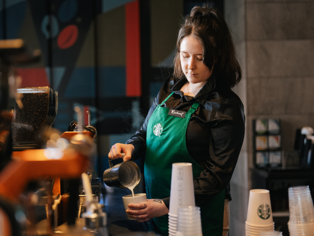 Une personne portant un tablier vert Starbucks se tient derrière le bar dans un magasin Starbucks et verse du lait dans une boisson.