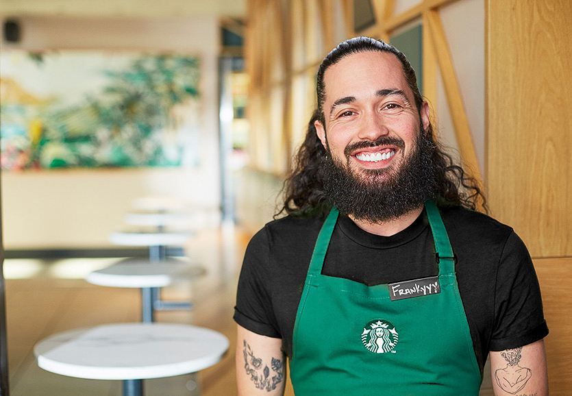 Une personne portant un tablier vert Starbucks sourit en regardant la caméra.