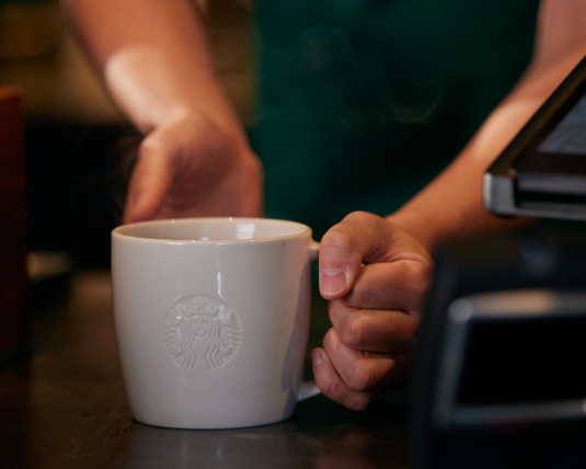 La main d’une barista Starbucks qui porte un tablier vert fait glisser une tasse en céramique blanche sur le comptoir d’un café.