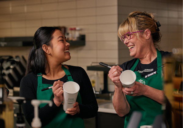 Une barista Starbucks et sa gérante rigolent en écrivant un message sur des tasses.