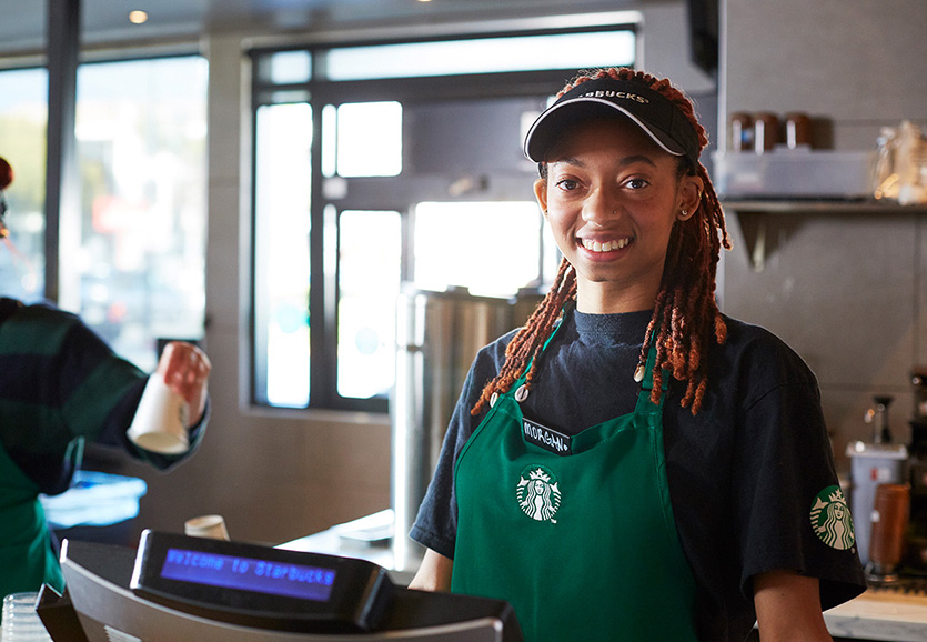 A Starbucks employee behind the counter in a store smiles while wearing a green apron and Starbucks visor.