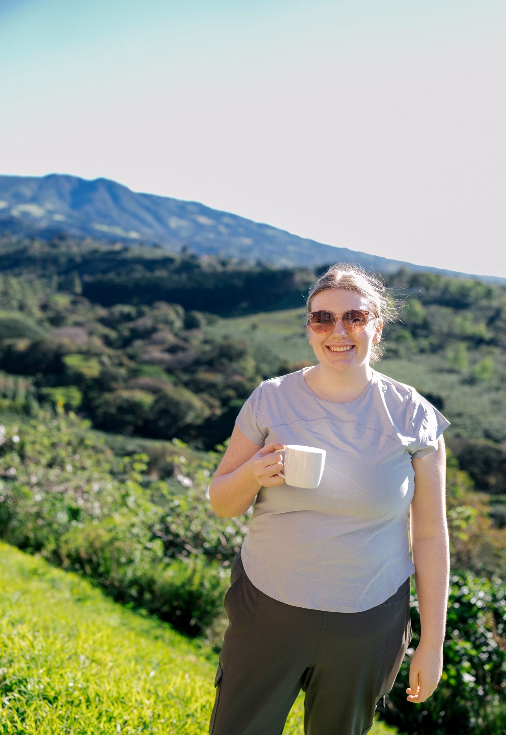 A person standing with a coffee farm in the background smiles into the camera while holding a cup of coffee.