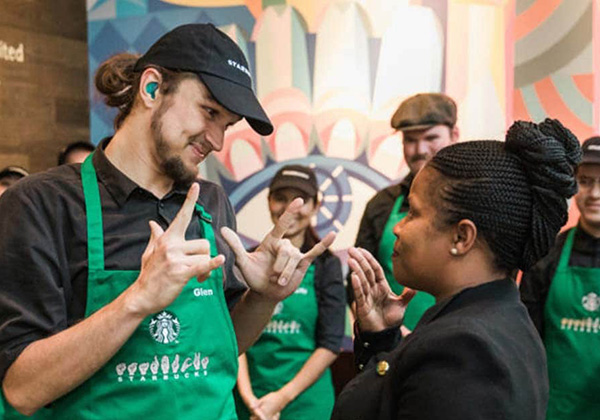 Two individuals wearing Starbucks green aprons communicate with each other using sign language, surrounded by fellow employees.