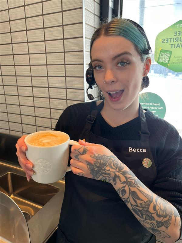 Starbucks employee wearing a black coffee master apron and a drive-thru headset, holds a white in-store cup with a latte and smiles at the camera.