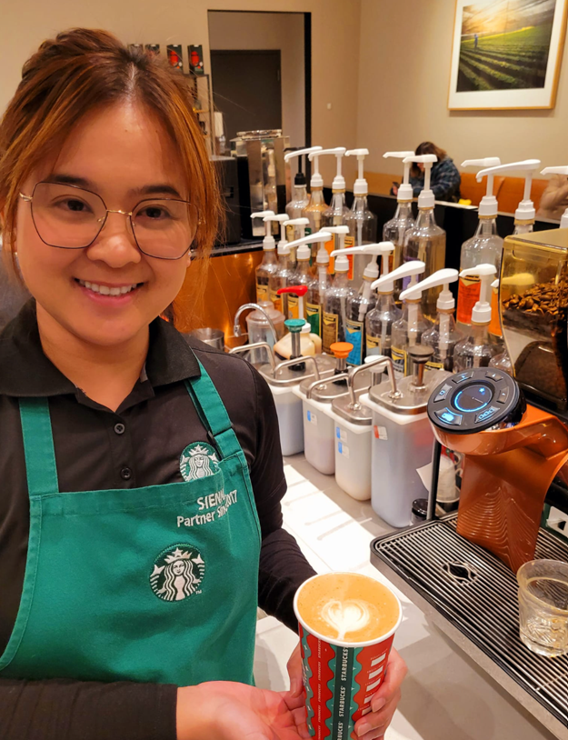 Starbucks employee wears glasses and a green Starbucks apron. She holds a coffee cup displaying latte art.