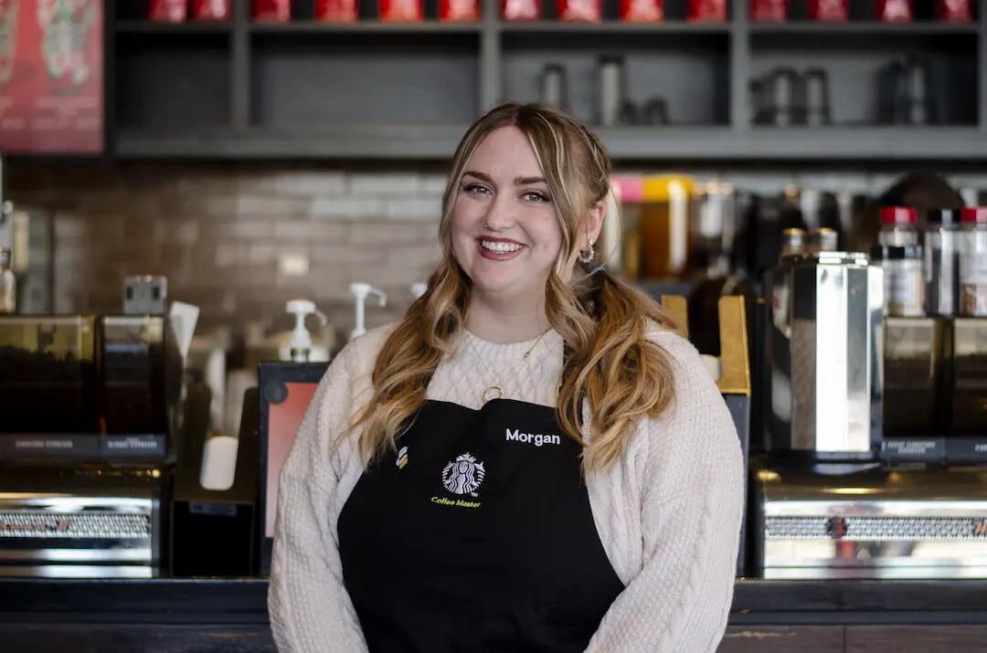 Starbucks employee wearing a black coffee master apron, has her hair in two pigtails and smiles warmly at the camera.