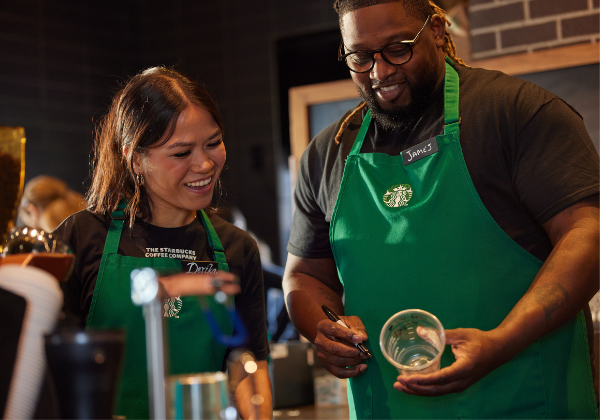 Two Starbucks baristas in a coffeehouse look at a cup that one has written on.