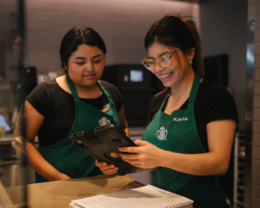 Two women Starbucks partners look at an iPad in the back of a Starbucks store. One points on the screen.