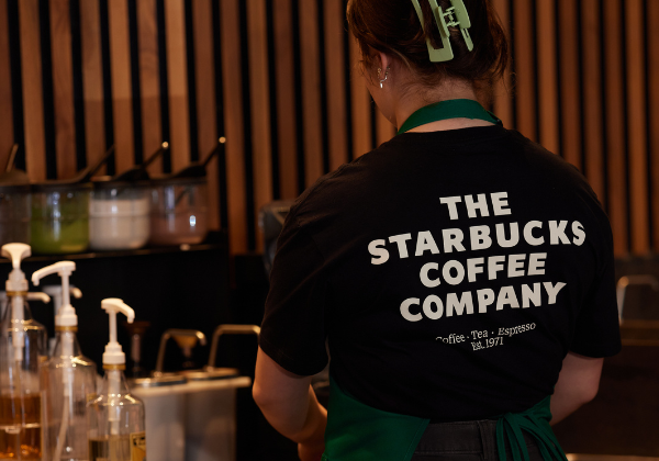 A barista at a Starbucks coffeehouse faces away from the camera. Her shirt reads, “The Starbucks Coffee Company”.