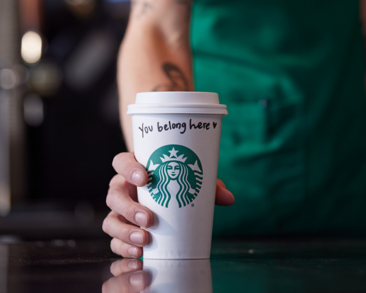 A hand from a Starbucks barista in a green apron pushes a white to-go-cup across the counter in a coffeehouse. On the cup it says, “you belong here”