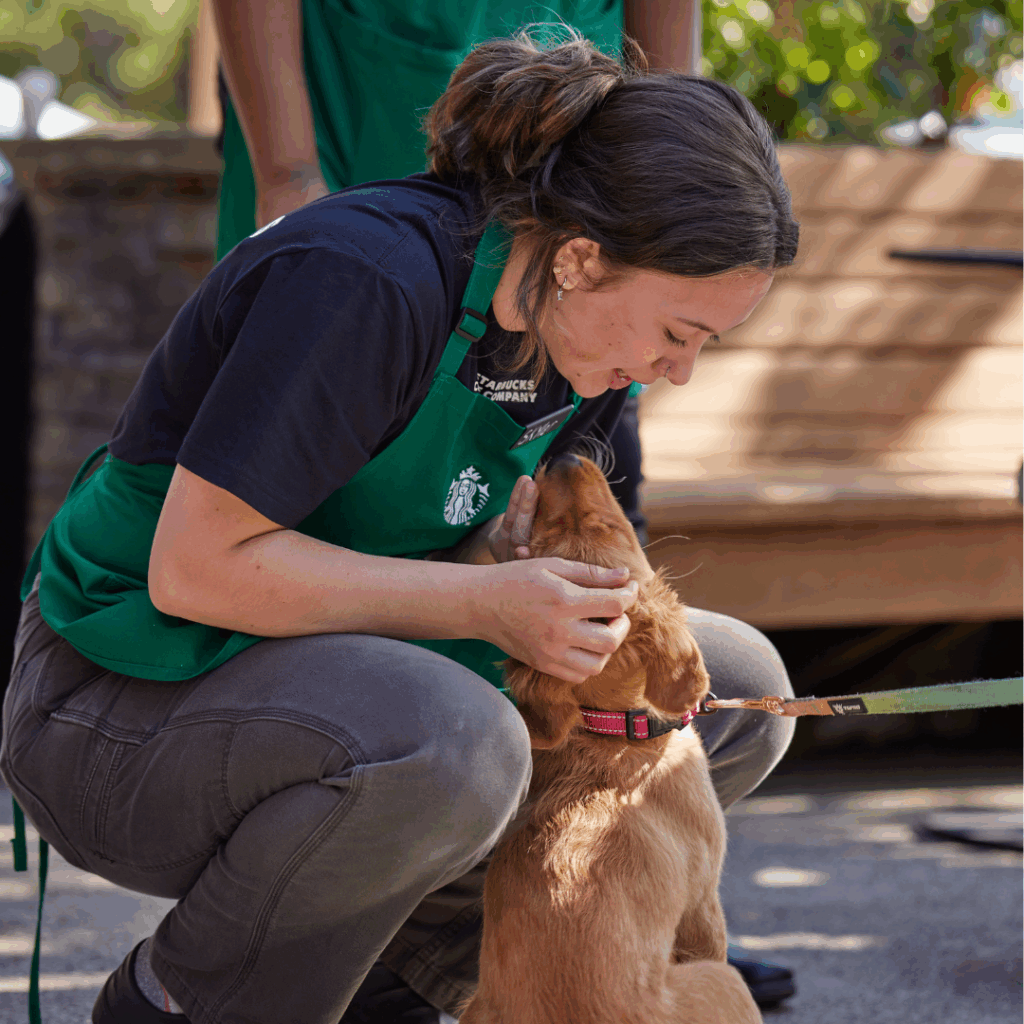 A Starbucks barista crouches down to pet a dog in front of a Starbucks coffeehouse.