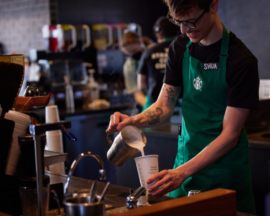 A Starbucks barista pours milk into a drink behind the bar at a Starbucks coffeehouse.