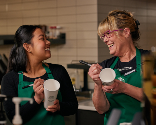 A Starbucks barista and her manager laugh together as they write on cups.
