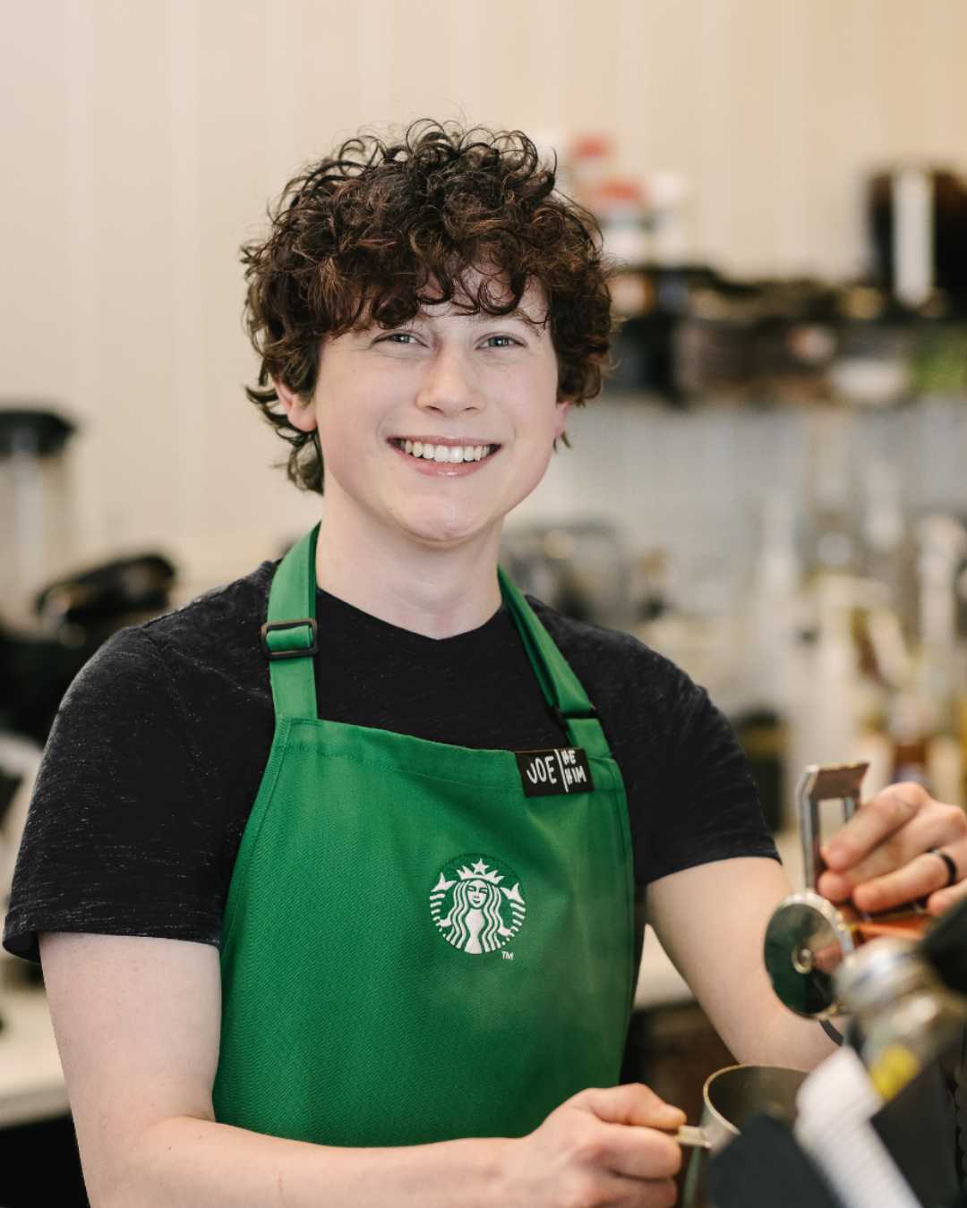 A person wearing a Starbucks green apron smiles directly into the camera.