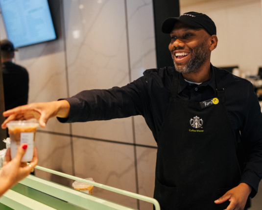 A person wearing a Starbucks Coffee Master black apron smiles as they hand a coffee to a customer over the counter while working at Starbucks.