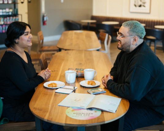 Two individuals working at a Starbucks coffeehouse sit across from each other and sip from coffee cups while in conversation.