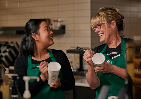 A Starbucks barista and her manager laugh together as they write on cups.