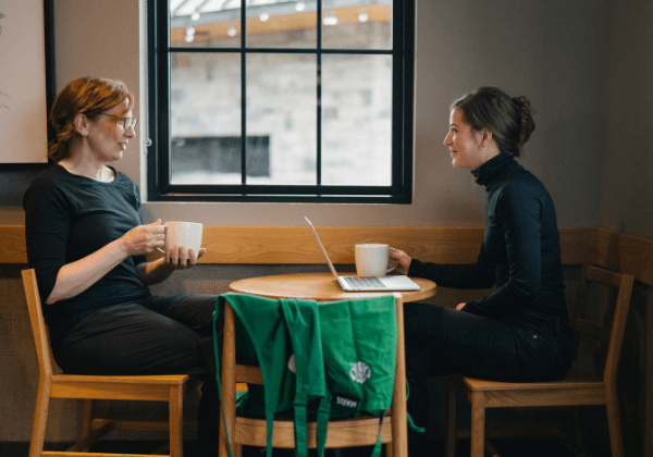 Two Starbucks partners sit in a coffeehouse at a round table. They are chatting with each other while one has their laptop open.